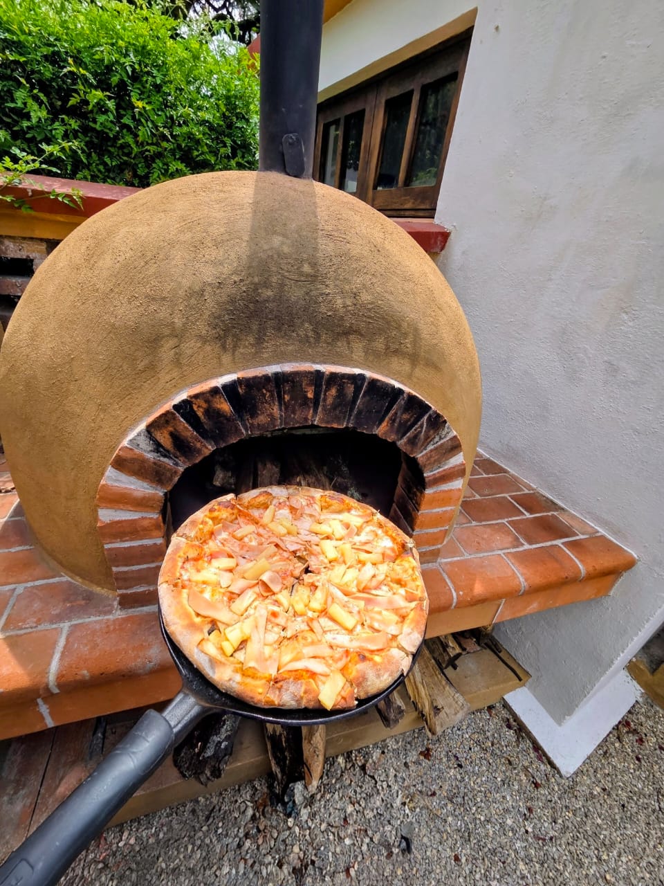 Round pizza with pineapple chunks and ham cooking in a domed outdoor clay oven, set on a brick ledge beside a white wall and chimney pipe.