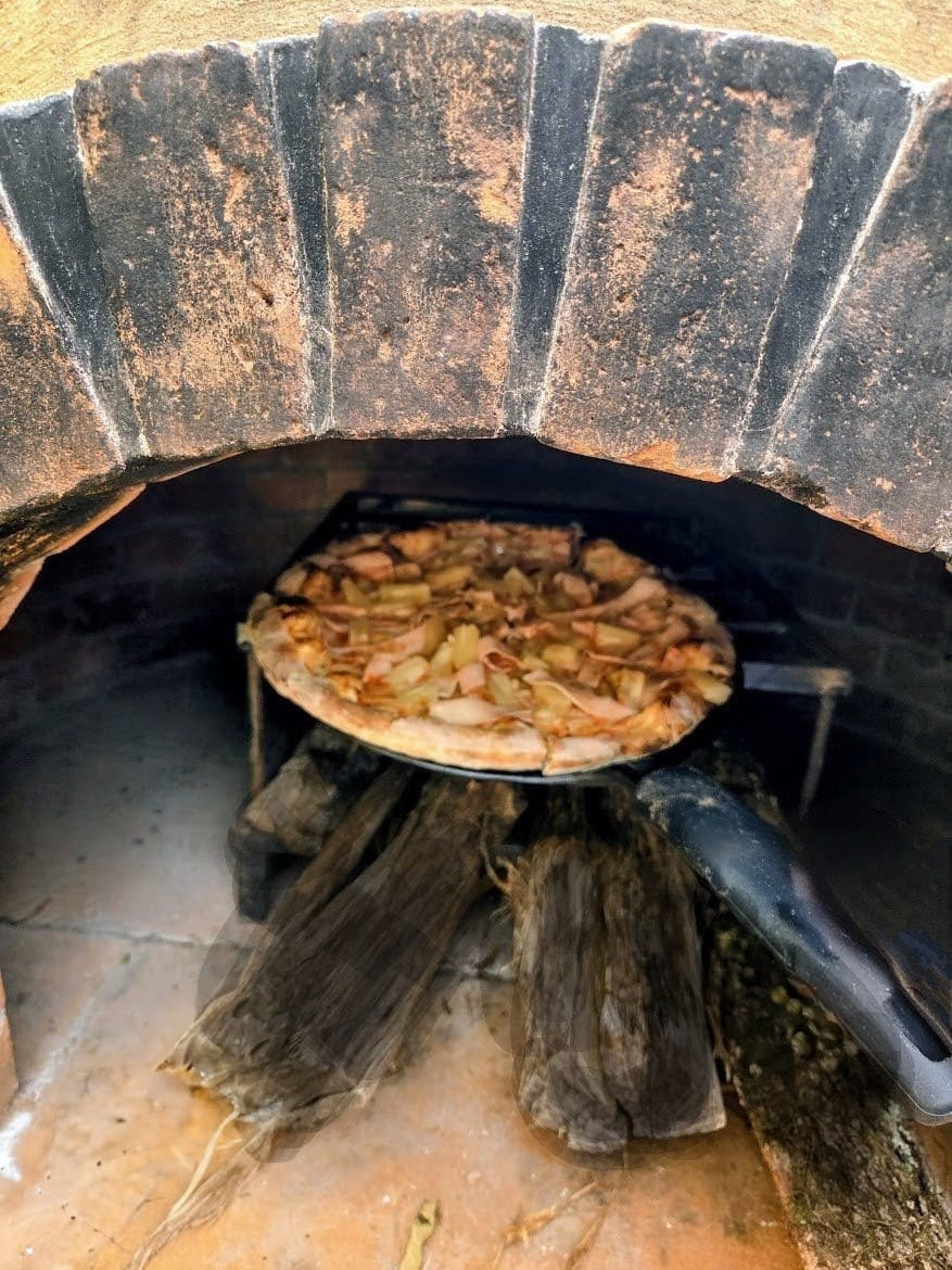 Pizza baking in a brick wood-fired oven on a round baking peel, with logs below.